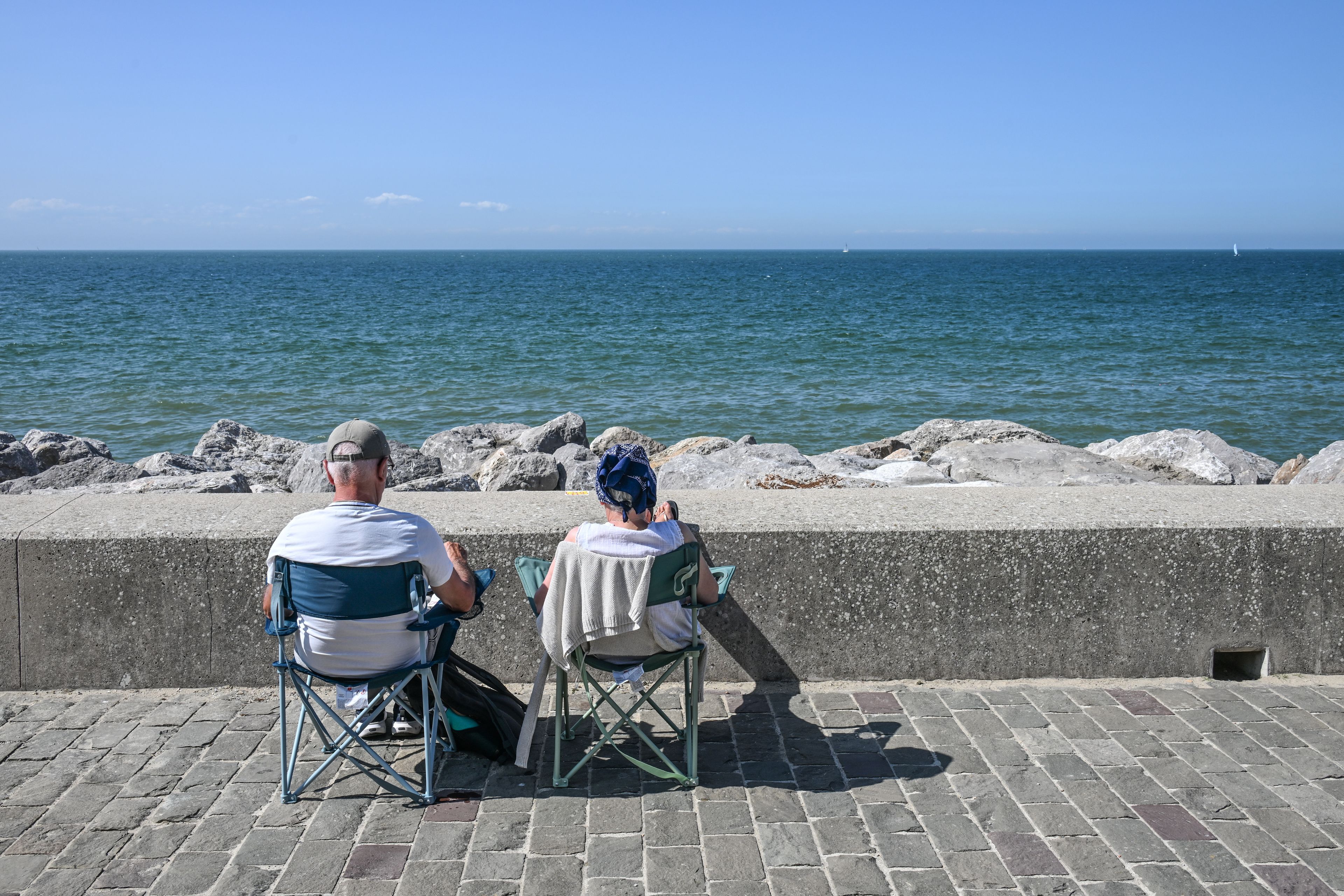 Dos personas sentadas en sillas mirando al mar, con una costa rocosa y cielo despejado.