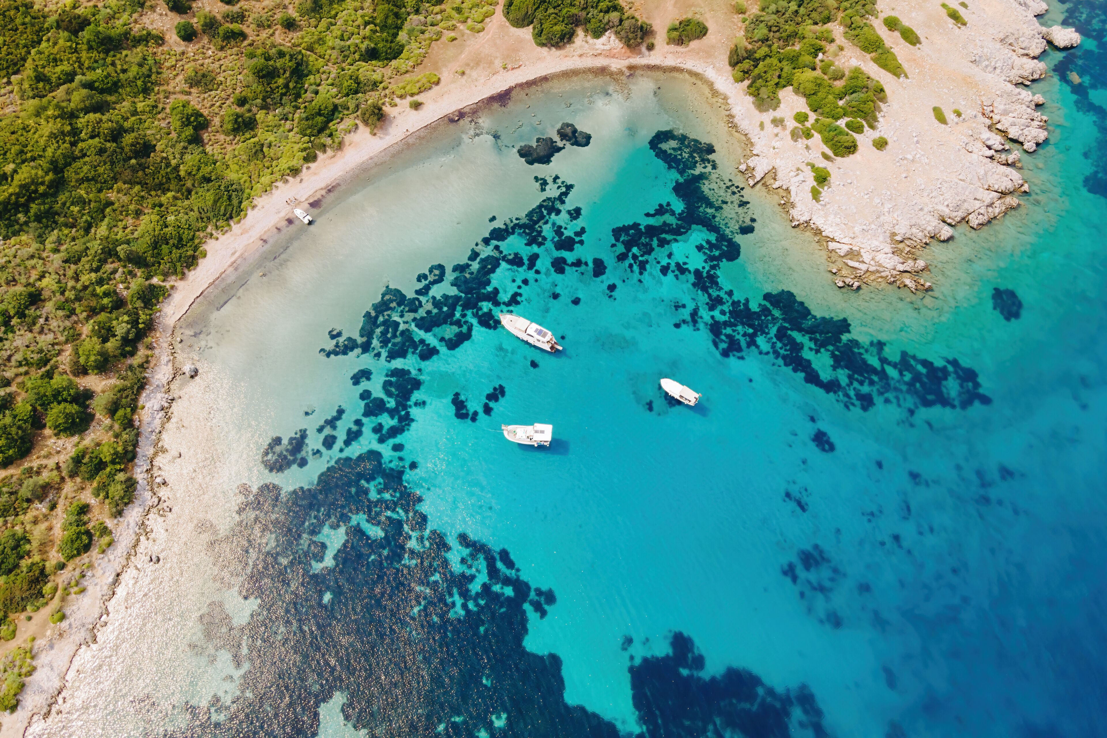 An aerial view of a turquoise coastal area with three boats anchored near the shore, surrounded by lush greenery and rocky outcrops.