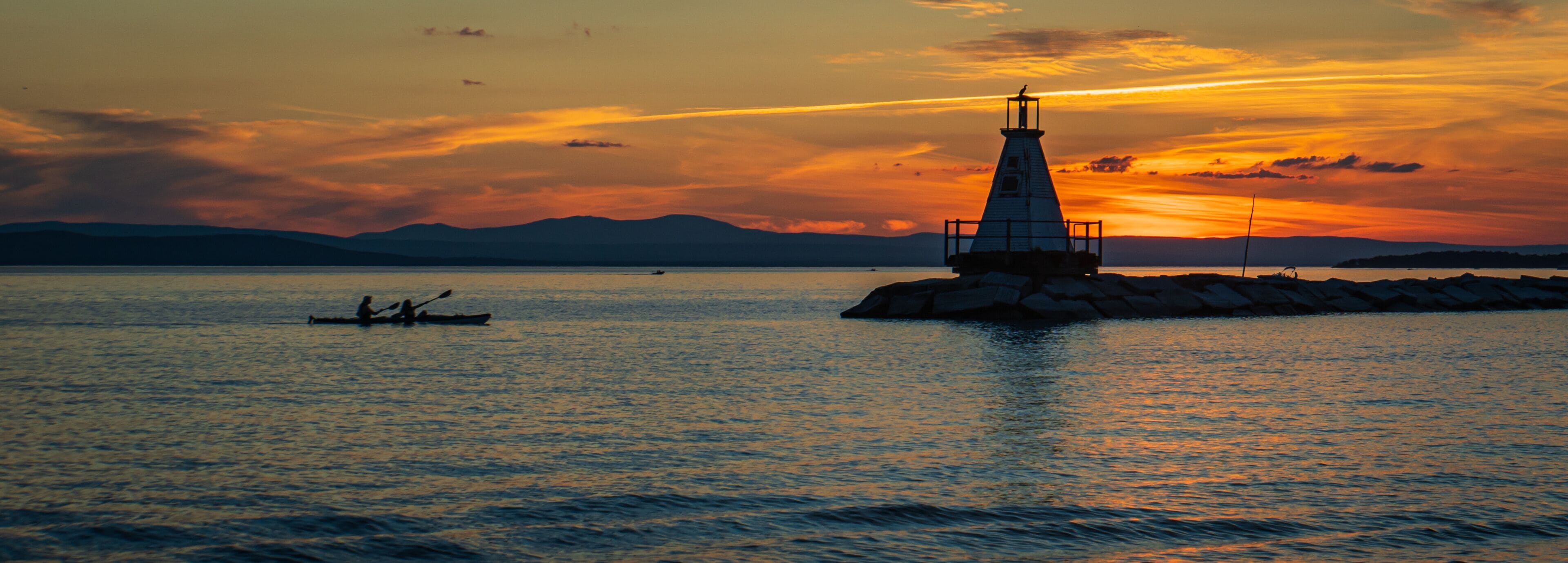 banner photo of kayakers approaching the Lake Champlain breakwater's lighthouse with a sea bird perched on the top at sunset 
