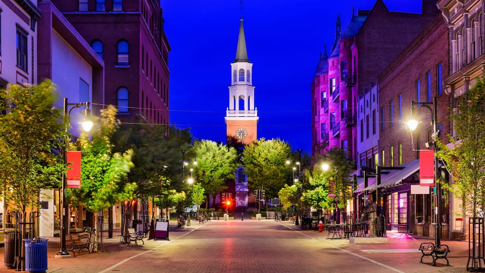 A historic town square with a prominent clock tower, lined with trees and street lamps.
