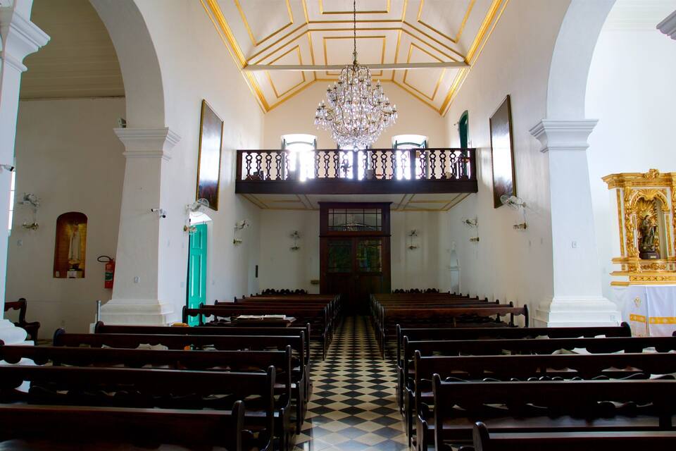 Interior de igreja com bancos de madeira, piso xadrez, lustre e uma estátua em um altar.