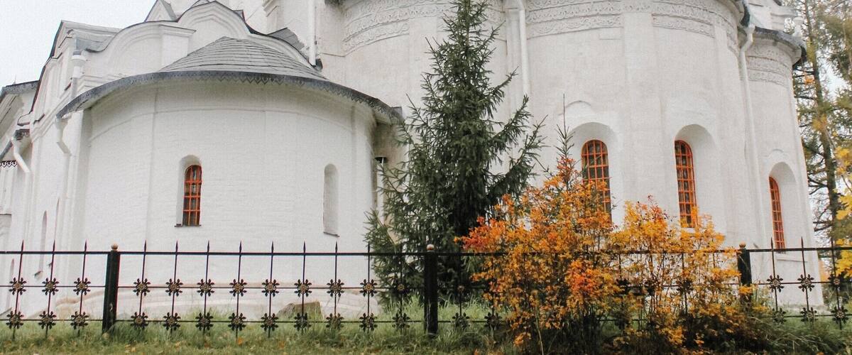 A white church with golden domes and a cross on top, surrounded by autumn leaves and a fence.