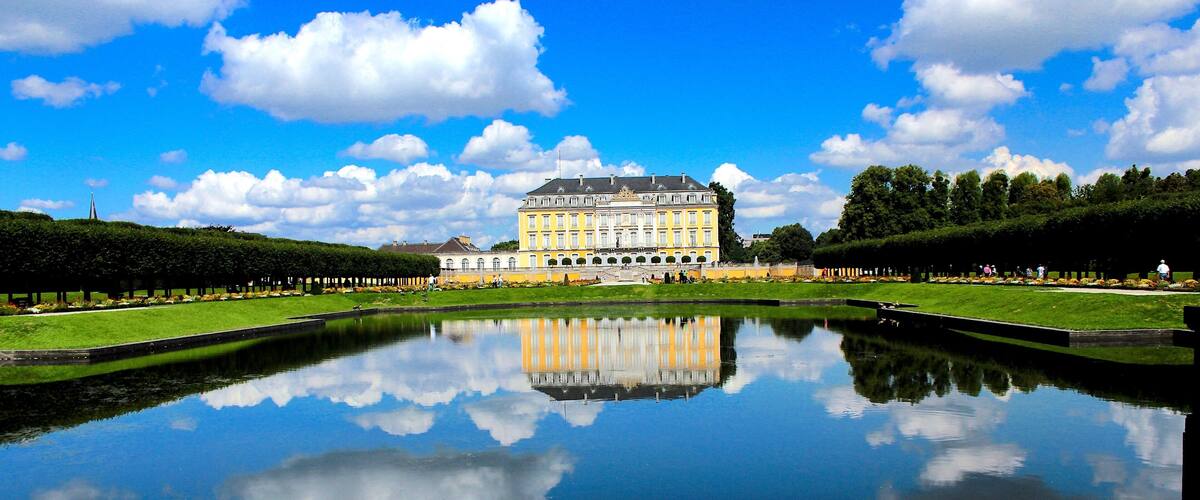 A large palace with a reflection in a pond, surrounded by manicured gardens and a clear blue sky.