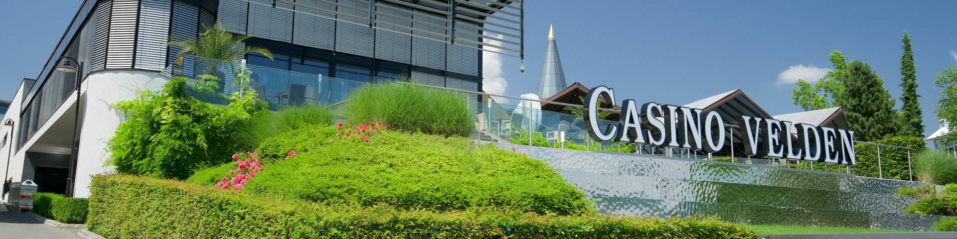 A modern building with a large sign reading "CASINO VELDEN" and a landscaped area with bushes and flowers.