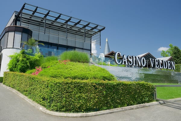 Ein modernes Gebäude mit einer großen Schrifttafel, auf der „CASINO VELDEN“ steht, und einer angelegten Grünfläche mit Büschen und Blumen.