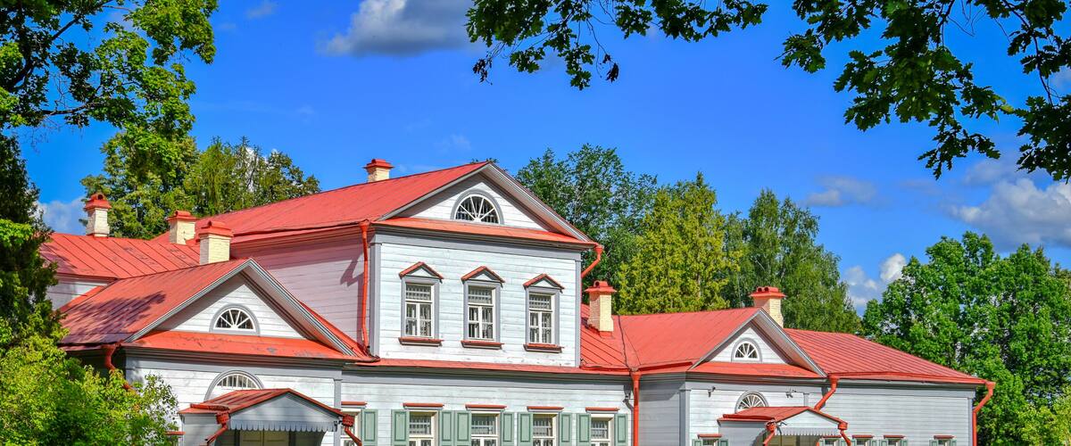 A large, two-story house with a red roof and green shutters, surrounded by lush greenery and trees.