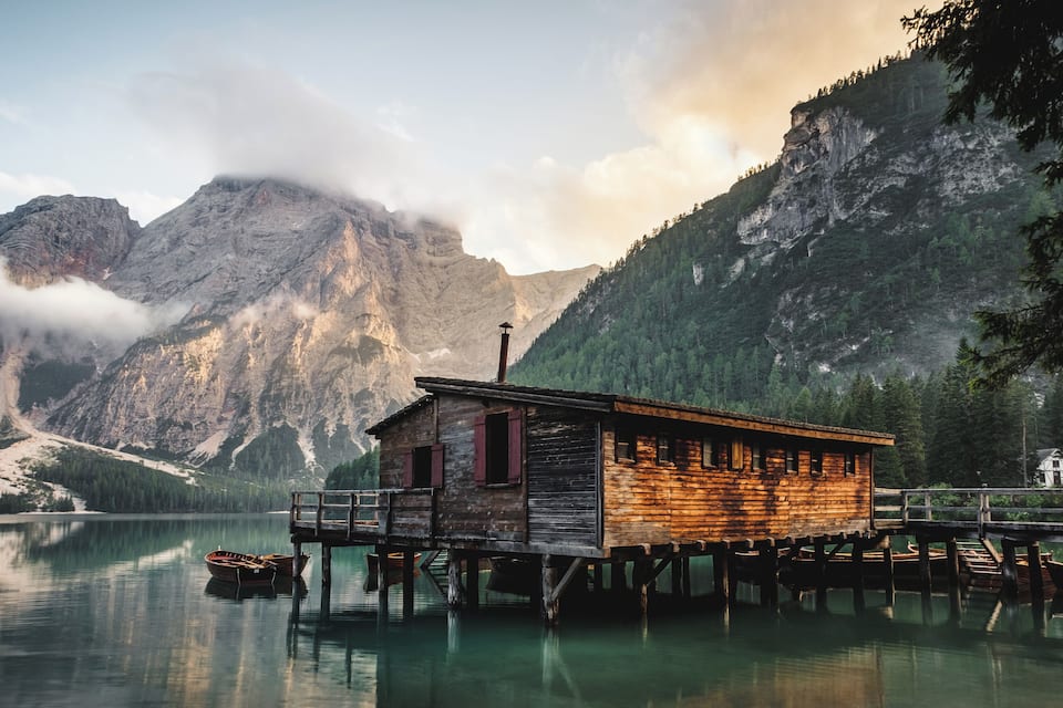 A wooden cabin on stilts by a calm lake, surrounded by mountains and trees.