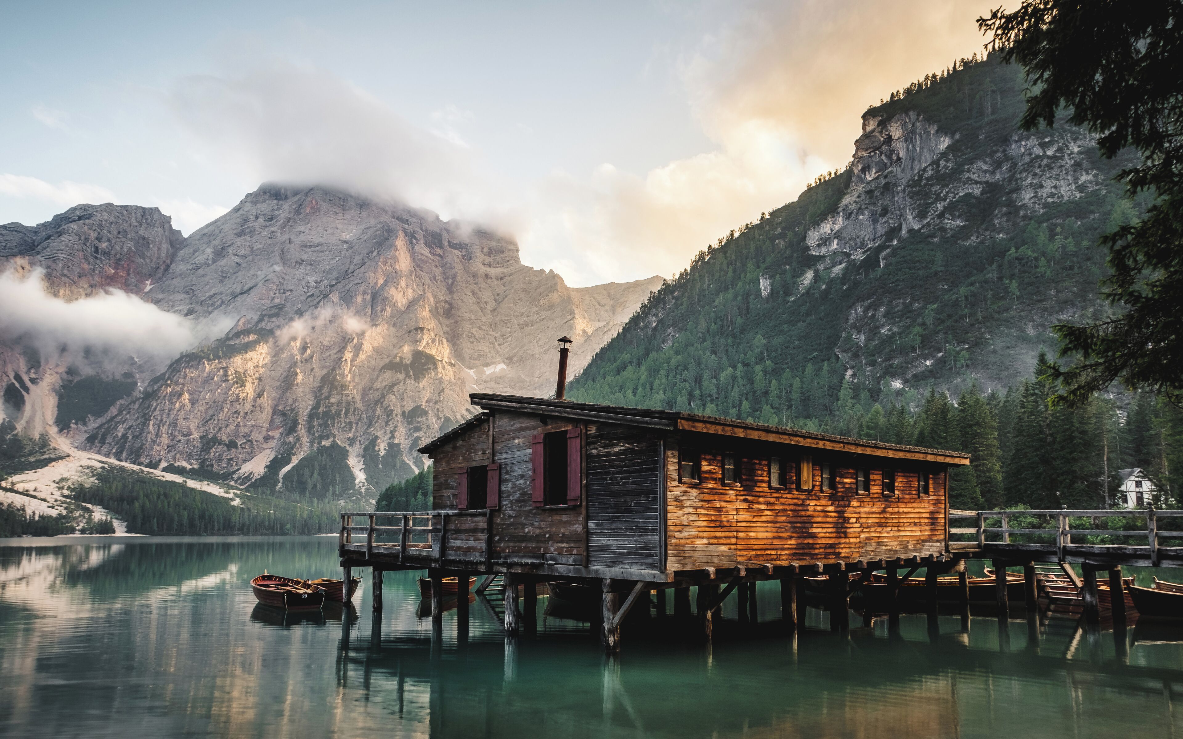 A wooden cabin on stilts by a calm lake, surrounded by mountains and trees.