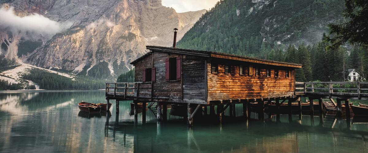 A wooden cabin on stilts by a calm lake, surrounded by mountains and trees.
