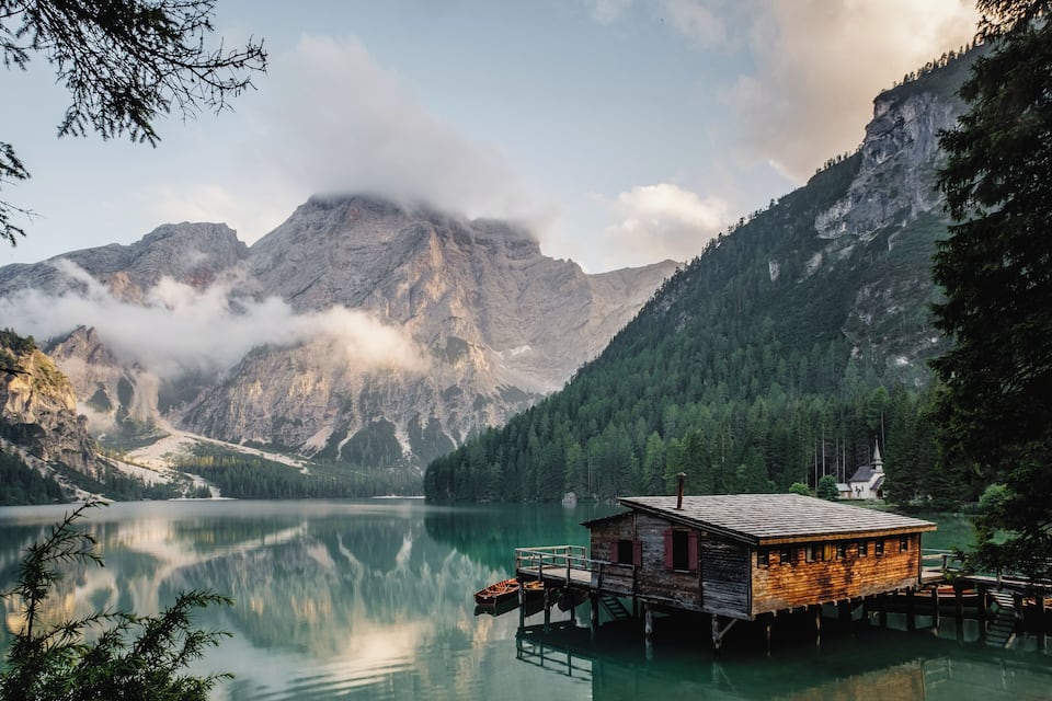 A wooden cabin on stilts over a calm lake, surrounded by mountains and dense forest.