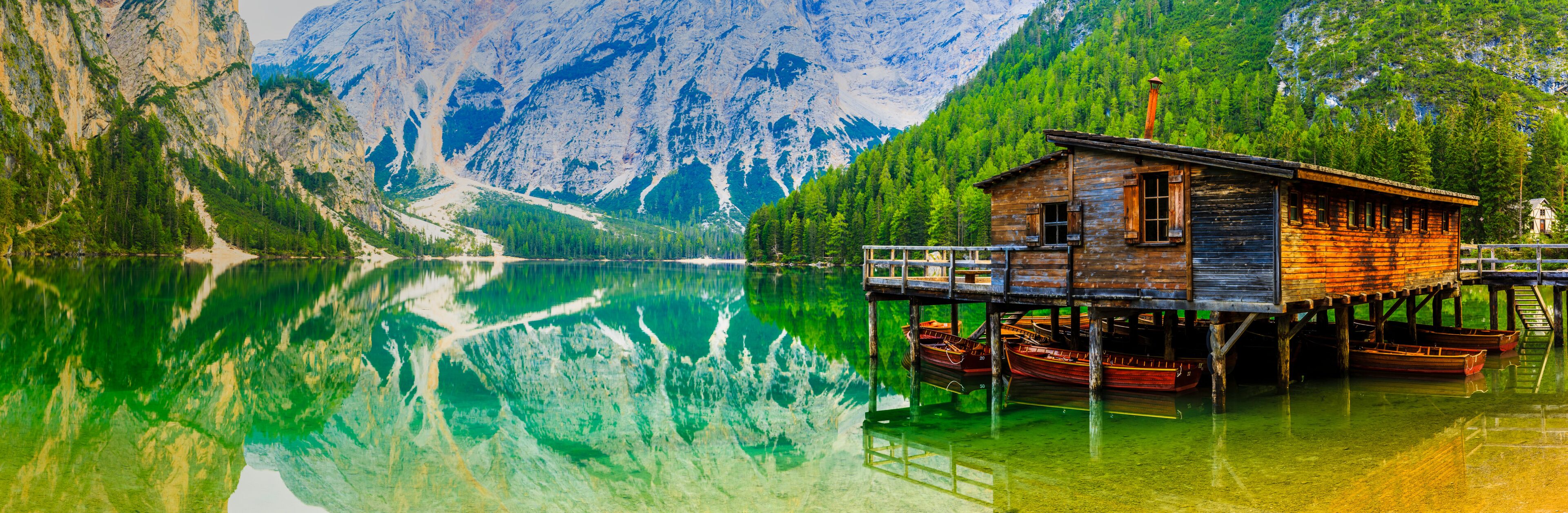 A wooden cabin on stilts by a clear lake with mountains in the background.