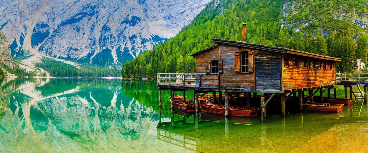 A wooden cabin on stilts by a clear lake with mountains in the background.