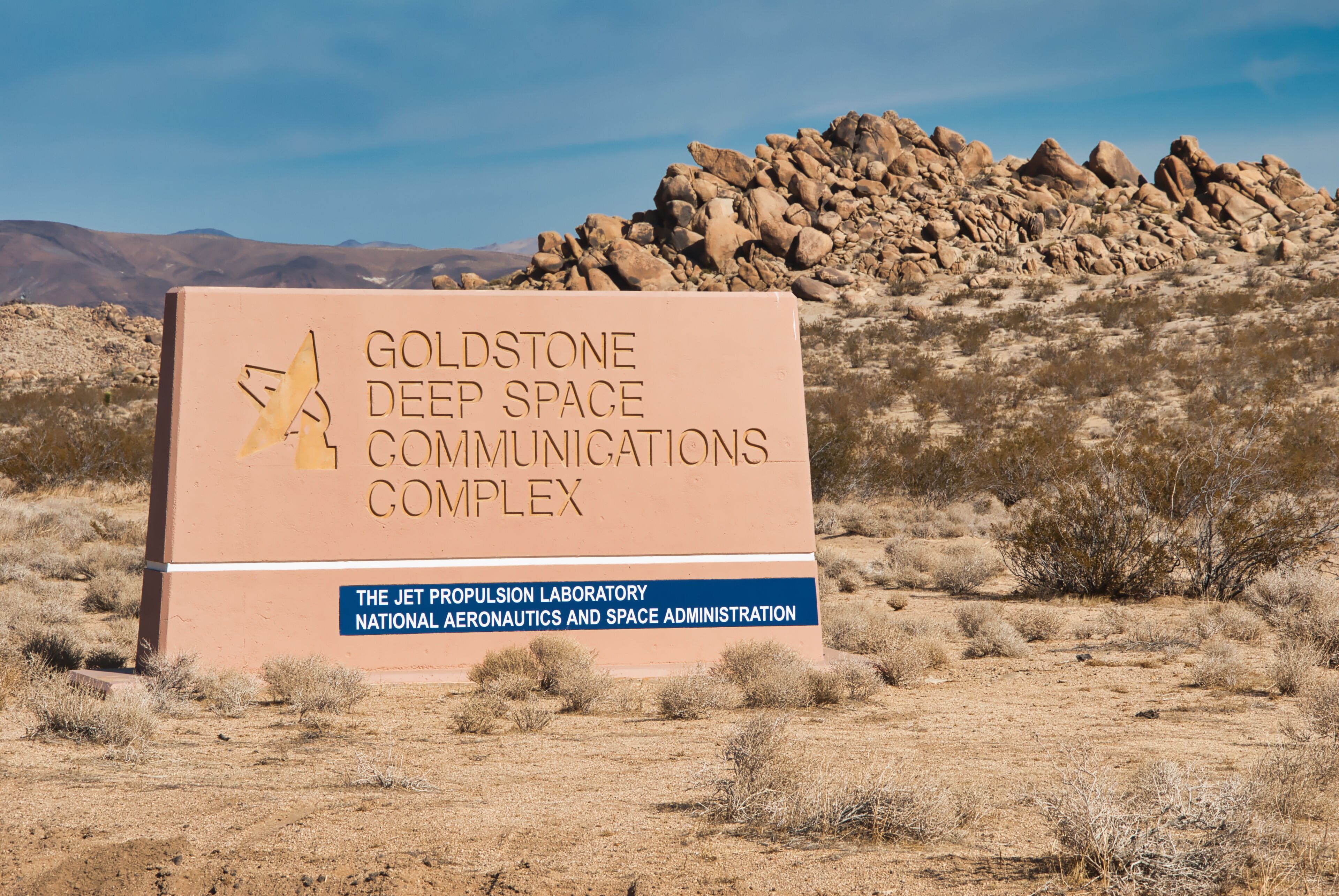 A sign for the Goldstone Deep Space Communications Complex with a rocky hill in the background.