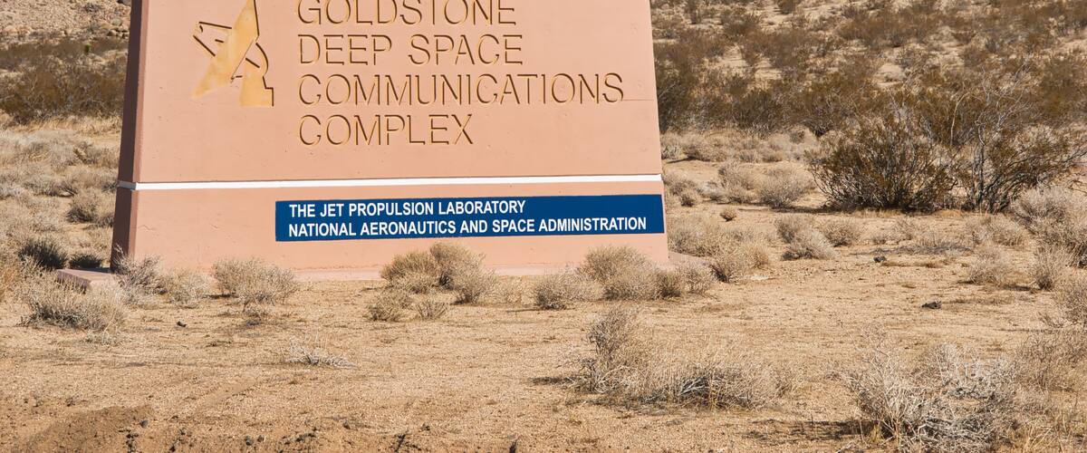 A sign for the Goldstone Deep Space Communications Complex with a rocky hill in the background.