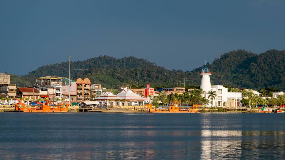 A coastal town with a prominent lighthouse, a harbor with boats, and a backdrop of green hills.