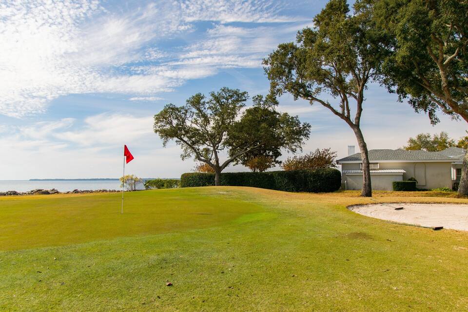 Un campo de golf con una bandera roja, árboles, una casa y una masa de agua al fondo.