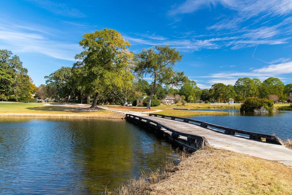 Un puente de madera sobre un lago tranquilo, rodeado de árboles y un cielo azul despejado.