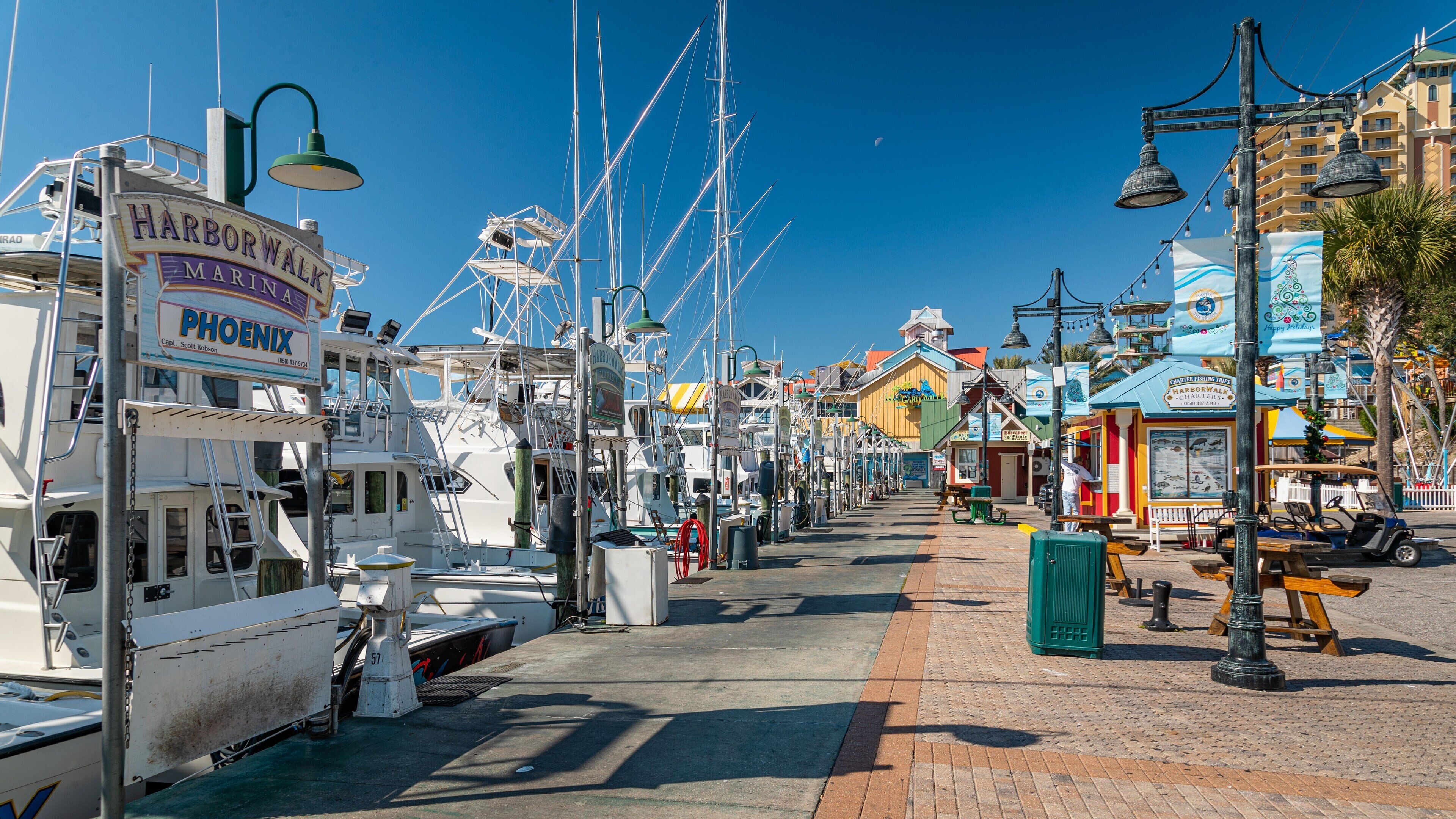 A marina with boats docked, a street with shops, and a clear blue sky.