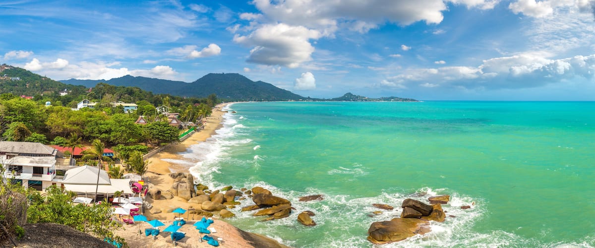 A coastal beach with turquoise waters, rocky outcrops, and a backdrop of mountains.
