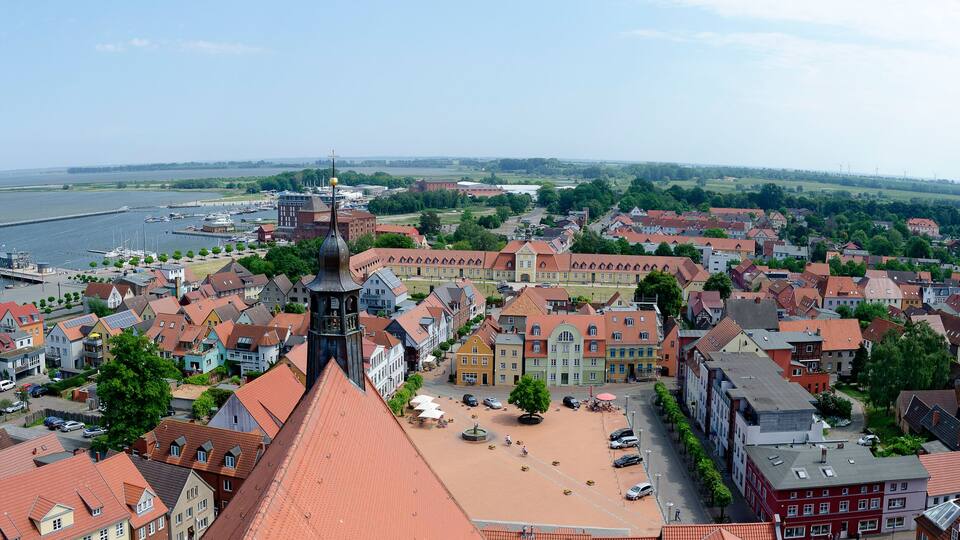 A panoramic view of a riverside town with historic buildings, a prominent church tower, and a marina with boats.