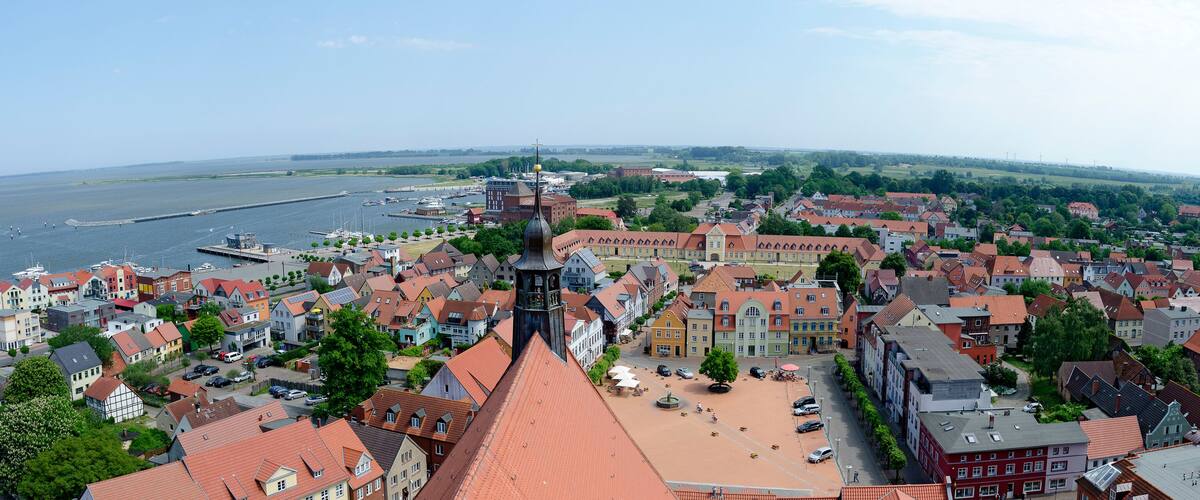 A panoramic view of a riverside town with historic buildings, a prominent church tower, and a marina with boats.