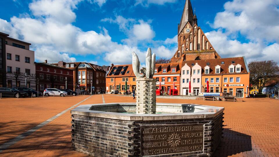 A historic church with a tall spire, a fountain with inscriptions, and a cobblestone square with buildings and parked cars.