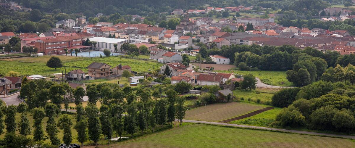 A town with red-roofed buildings, green fields, and a forested hill in the background.