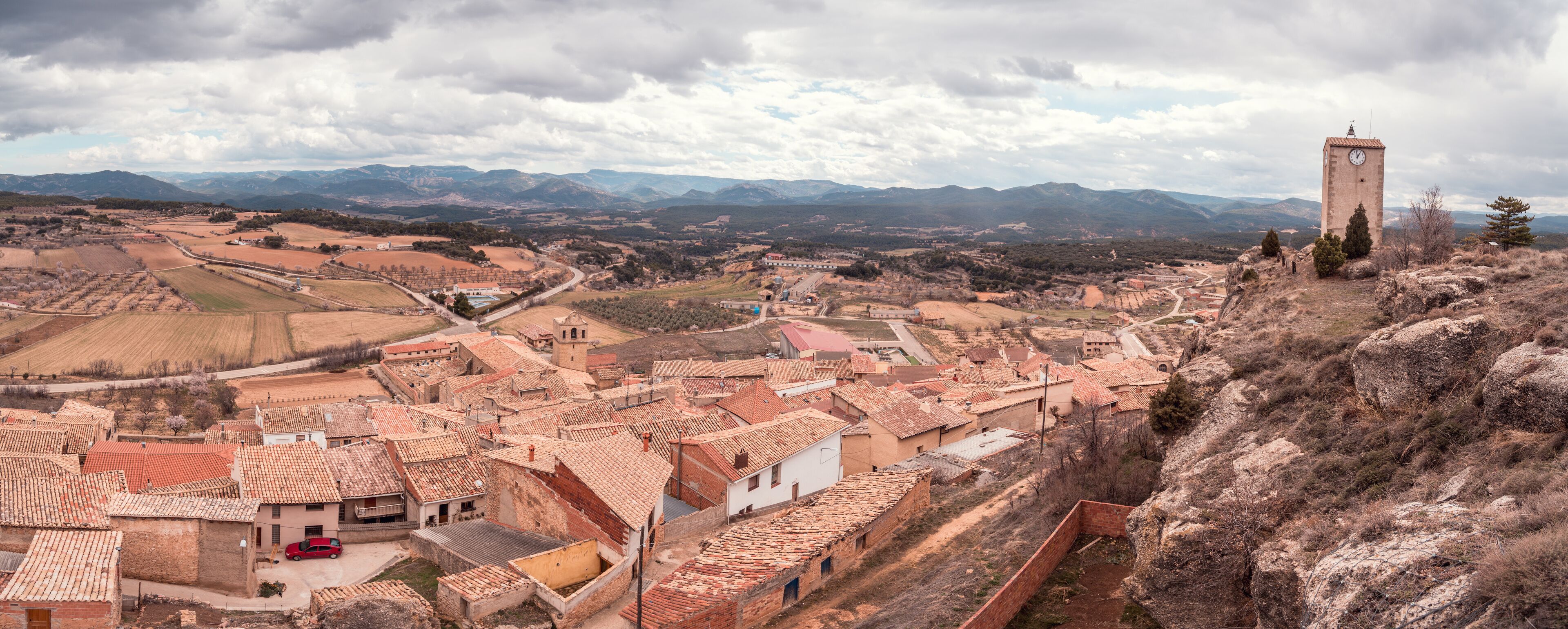 A town with red-tiled roofs, a prominent clock tower, and a mountainous backdrop.