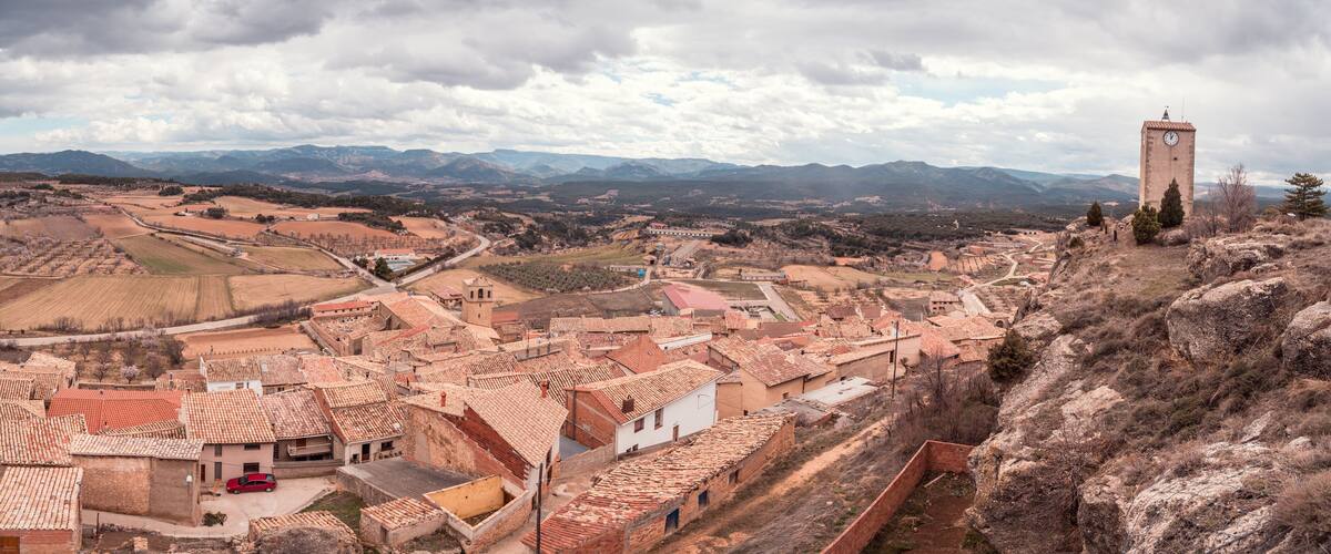 A town with red-tiled roofs, a prominent clock tower, and a mountainous backdrop.