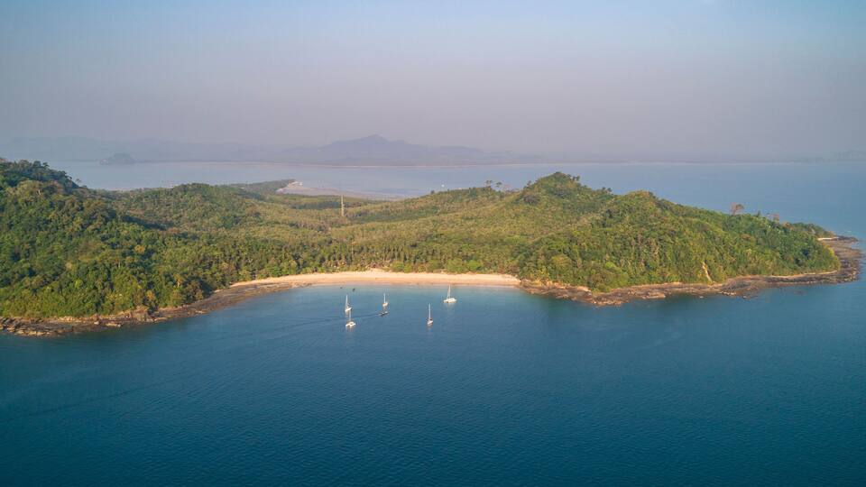 A coastal landscape with a sandy beach, dense green hills, and a calm sea with sailboats.