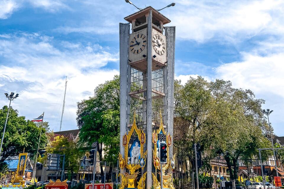 A clock tower with a golden statue base, surrounded by greenery and urban elements.