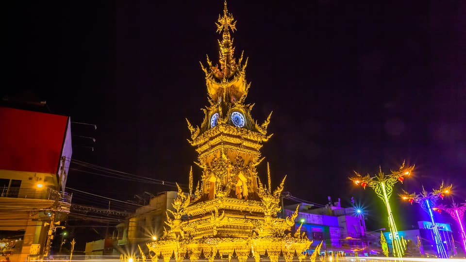 A brightly lit, ornate temple with multiple tiers and golden decorations, surrounded by a reflective water surface and colorful lights.