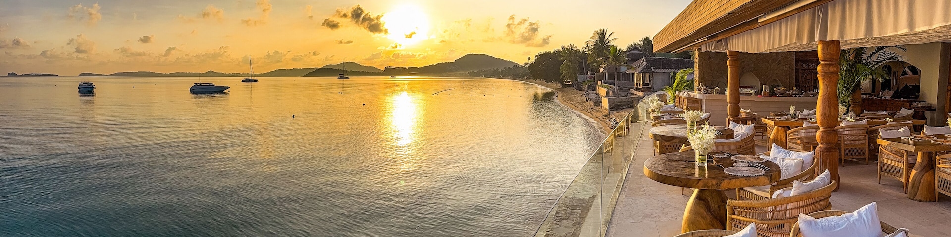 Una playa tranquila con un edificio de madera, muebles de mimbre y aguas calmadas que reflejan el atardecer.