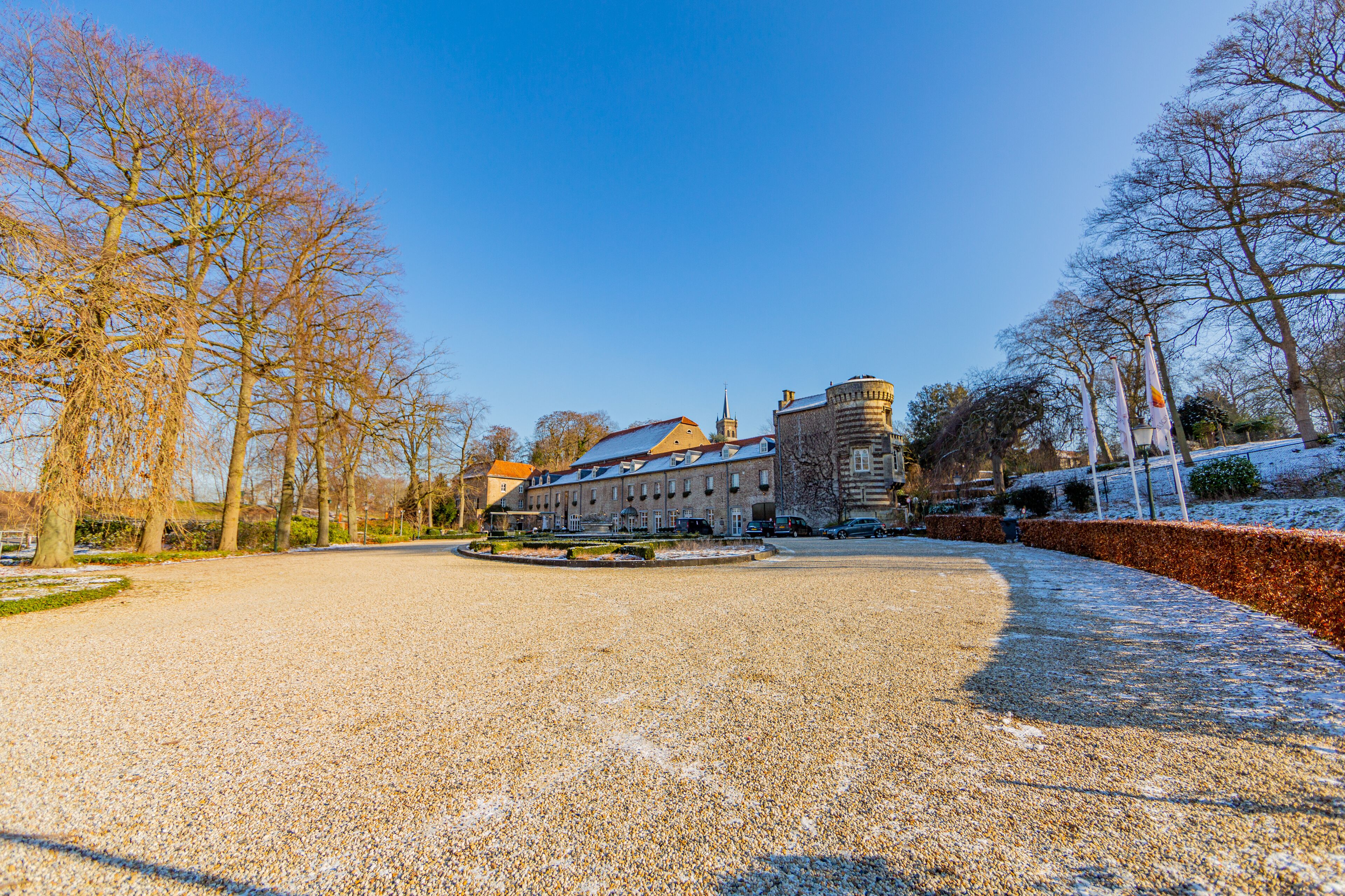 Esplanade of land surrounded by bare trees, shrubs with remnants of snow with Elsloo castle in the background, sunny winter day with a blue sky in Elsloo, South Limburg, Netherlands