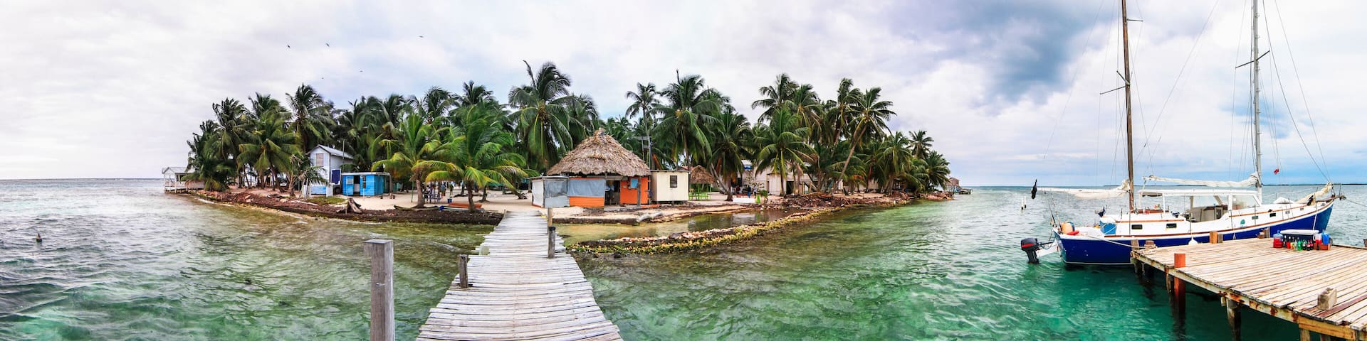 A wooden pier leading to a small island with thatched-roof huts and palm trees.