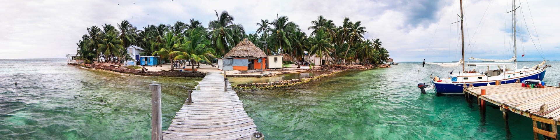 Eine Holzstege führt zu einer kleinen Insel mit Lehmhütten und Palmen.