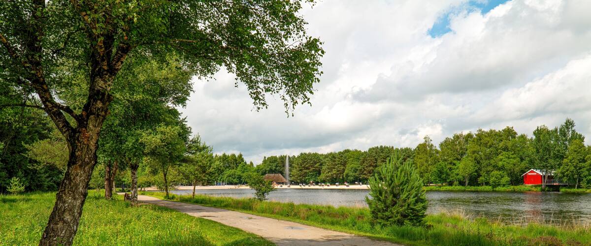 A park with a dirt path, a lake, a red barn, and a thatched-roof building.