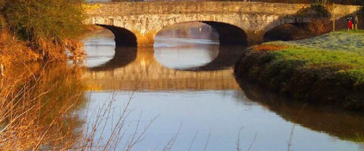 Un pont de pierre enjambe une rivière paisible dont les eaux reflètent le ciel, le tout étant entouré d’arbres et bordé d’une rive herbeuse.
