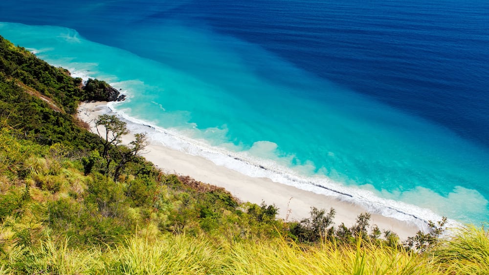 A coastal landscape with a sandy beach, clear turquoise waters, and lush greenery.