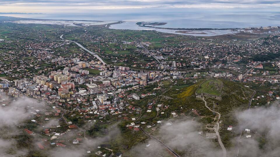 An aerial view of a coastal city with a dense urban center, surrounded by green fields and a coastline.