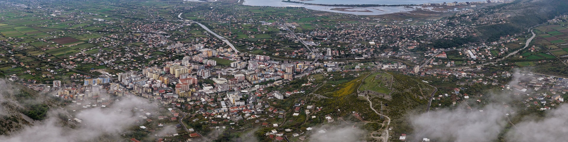 Wide aerial panorama showing Lezhë, Lezhë Castle, the Drin River, and Shëngjin stretching to the Adriatic Sea under cloudy skies.