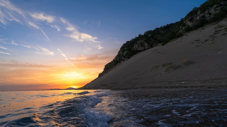 A beach at sunset with waves, a sandy shore, and a cliff.
