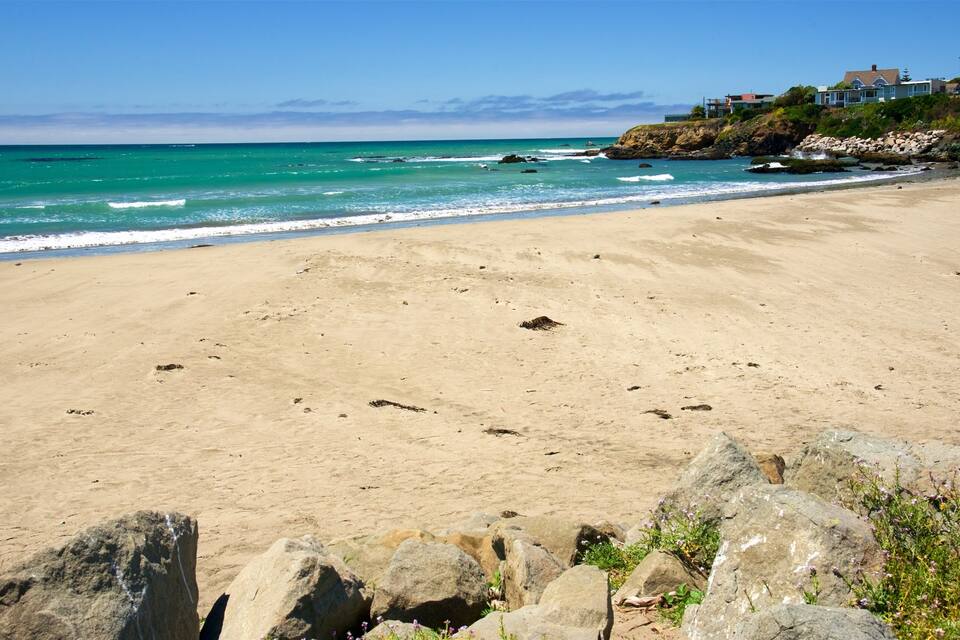 A sandy beach with waves, rocks, and houses on a cliff.