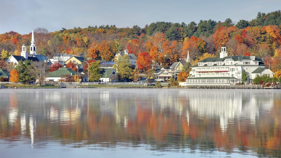 A lakeside town with autumn-colored trees, a church, and a large white building with a green roof.