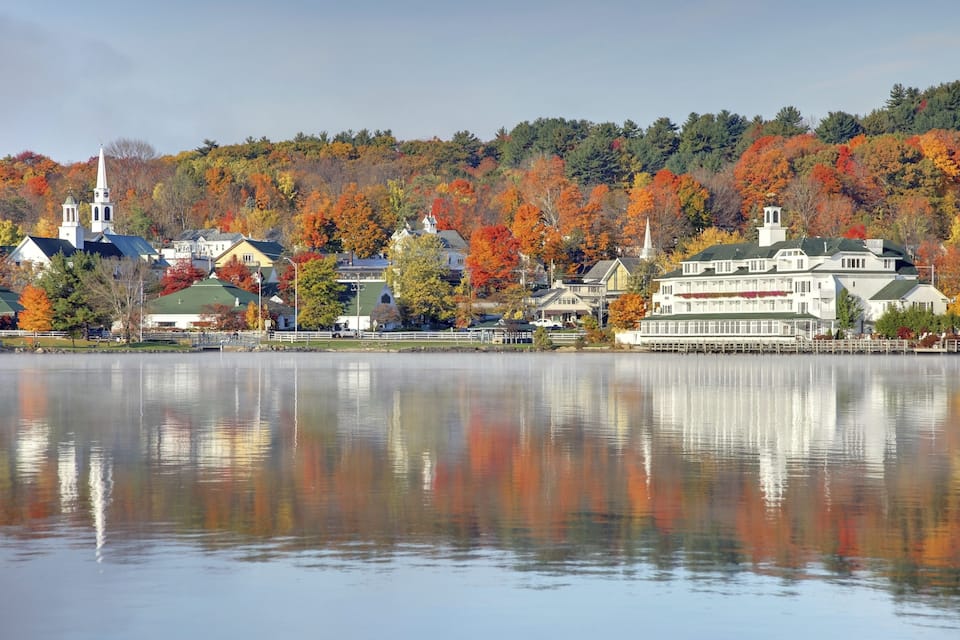 A lakeside town with autumn-colored trees, a church, and a large white building with a green roof.