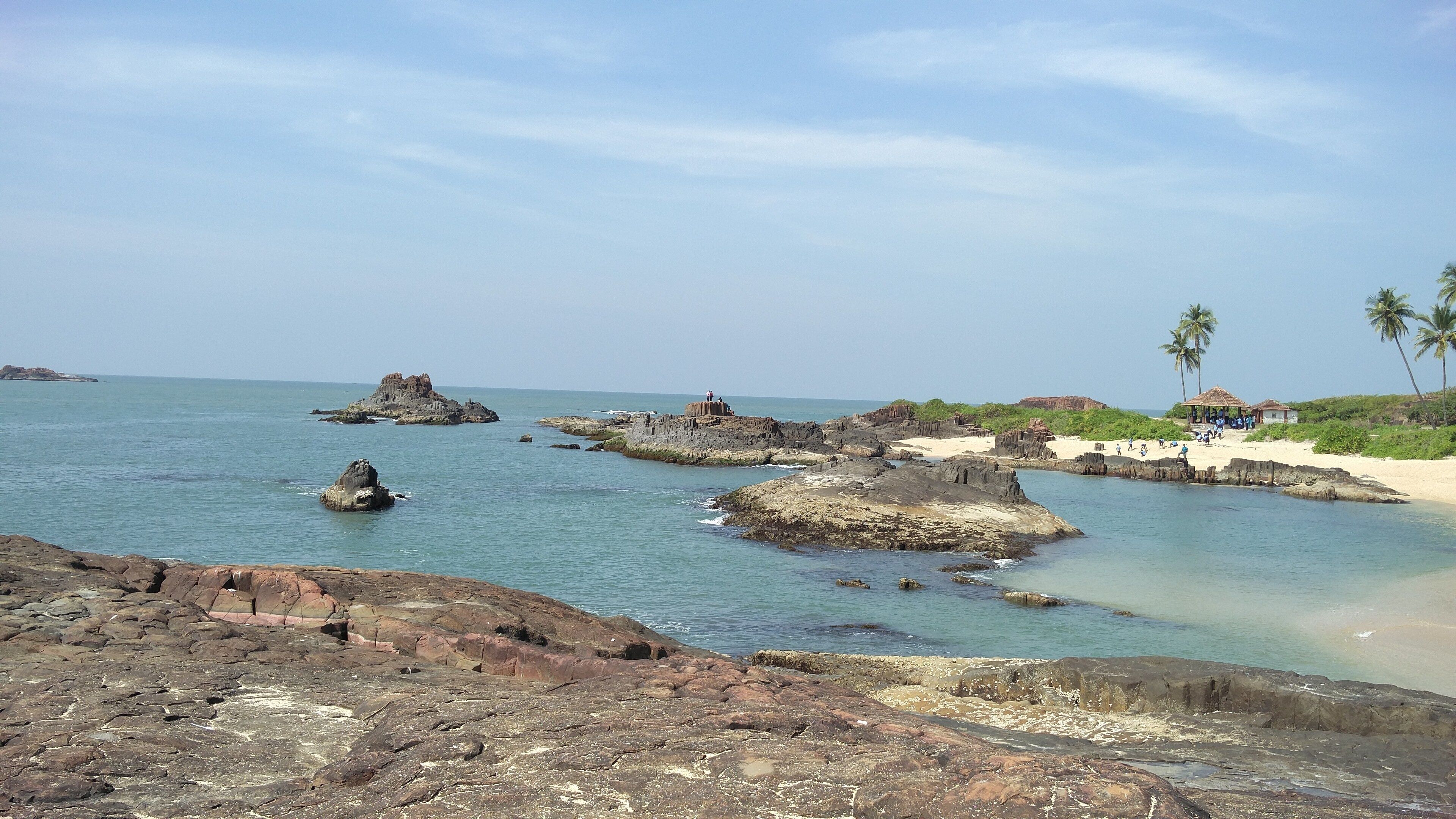 A rocky coastline with clear turquoise waters, palm trees, and a small beach with people.