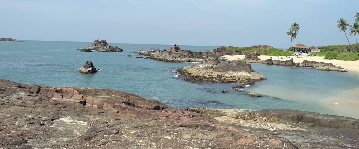A rocky coastline with clear turquoise waters, palm trees, and a small beach with people.