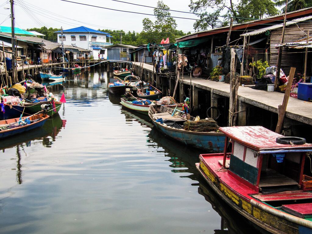 A narrow waterway with small boats docked along the sides, wooden houses, and a larger building in the background.