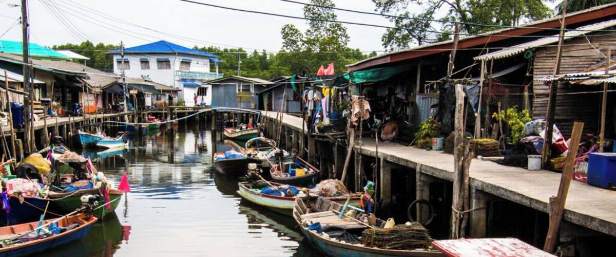 A narrow waterway with small boats docked along the sides, wooden houses, and a larger building in the background.