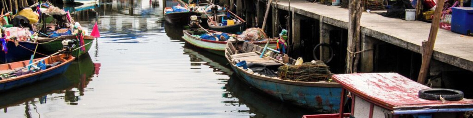 A narrow waterway with small boats docked along the sides, wooden houses, and a larger building in the background.